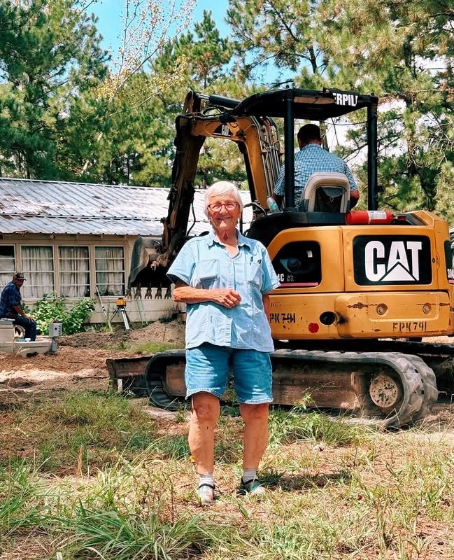Alma "Alligator Annie" Tippins smiling in front of a CAT mini excavator at a job site among pine trees