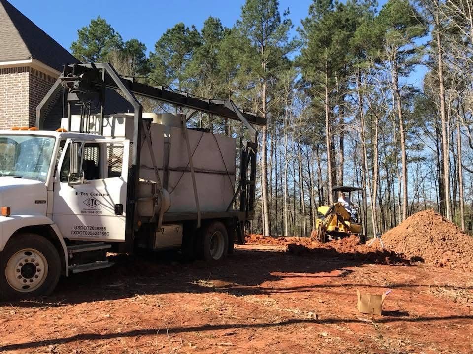 Concrete tank delivery truck and a small excavator at a red-clay job site among tall pine trees
