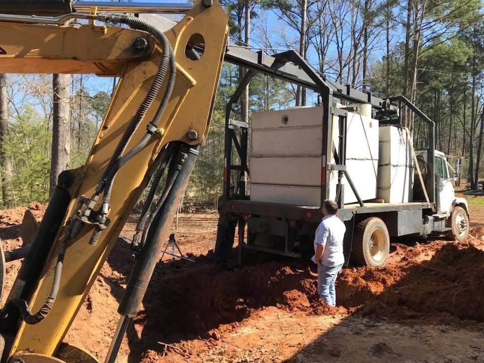 Excavator arm beside a boom truck carrying concrete tanks, with a worker standing in the trench